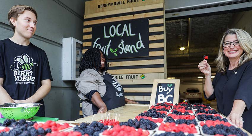Une femme tient une fraise devant un présentoir de fruits locaux.