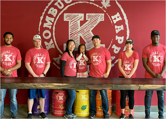 Des employés de Happy Belly Kombucha, en chandails rouges, debout derrière une table.