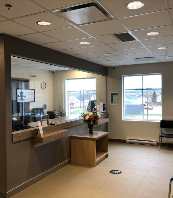 An image of a well-lit reception area with a counter, plexiglass barrier, floral arrangement, and seating.